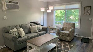 Un salon avec un canapé gris, des coussins à motifs, une table basse blanche, un fauteuil à motifs, un lampadaire et une fenêtre donnant sur un paysage verdoyant.
