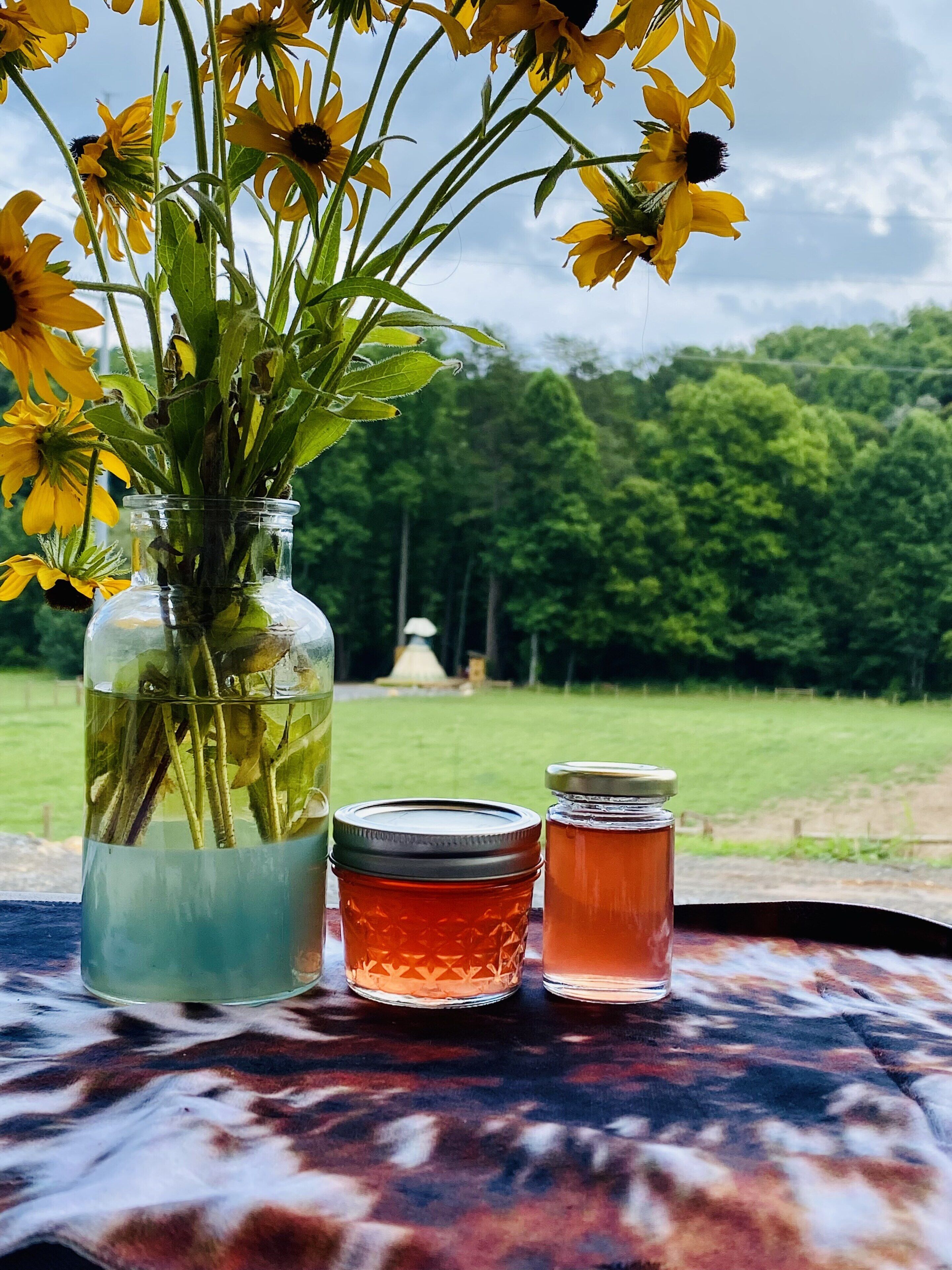 A table with a vase of yellow flowers, two jars of honey, and a view of a green landscape.