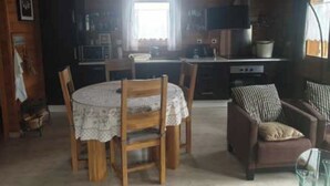 A cozy cabin-style room with a dining area featuring a round table and wooden chairs, a sofa with striped cushions, and a kitchenette with a stove and microwave. - Comfort chalet in the heart of Auvergne  (Maurines)