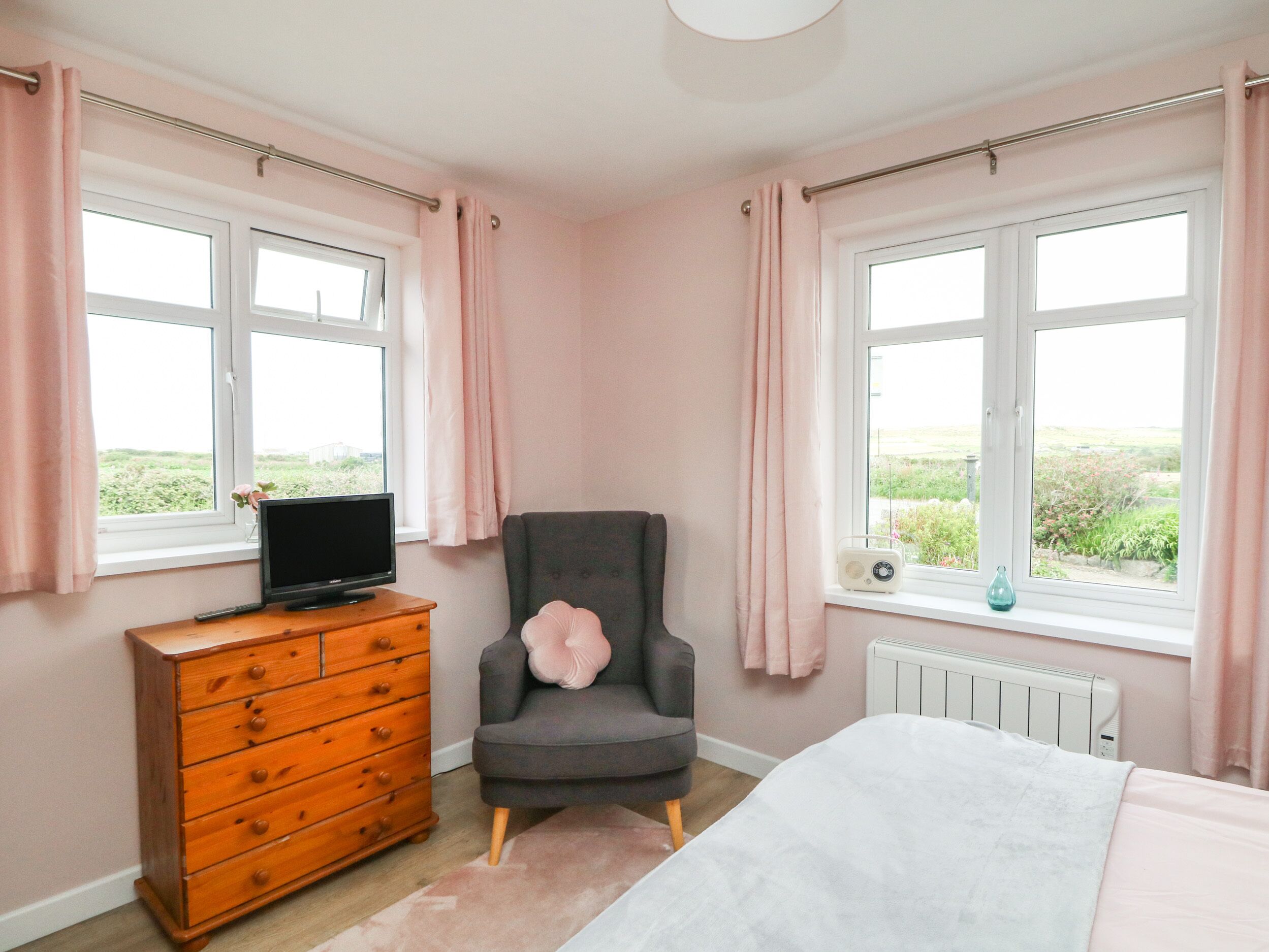 A bedroom with a bed, a dresser, a chair, a TV, and two windows with curtains.
