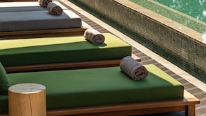 Un espace aménagé au bord de la piscine, avec des chaises longues rembourrées de couleur verte, un tabouret en bois et un grand parasol.
