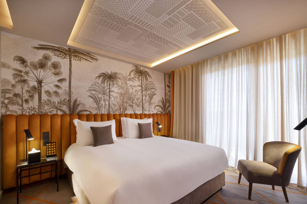 Una habitación de hotel con una cama grande, una mesita de noche con lámpara, una silla y un mural en la pared con palmeras.
