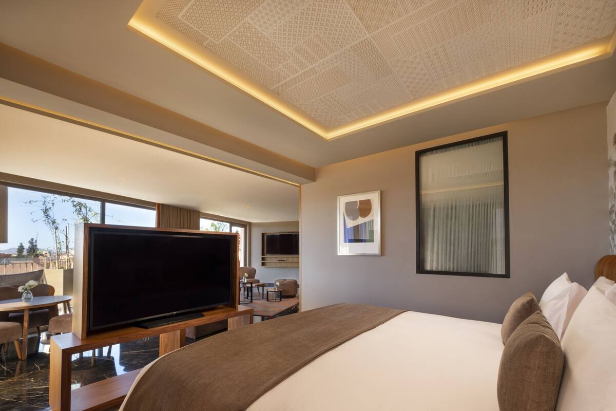 Una habitación de hotel moderna con una cama grande, un televisor de pantalla plana, una zona de comedor con mesa y sillas, y vistas al exterior.