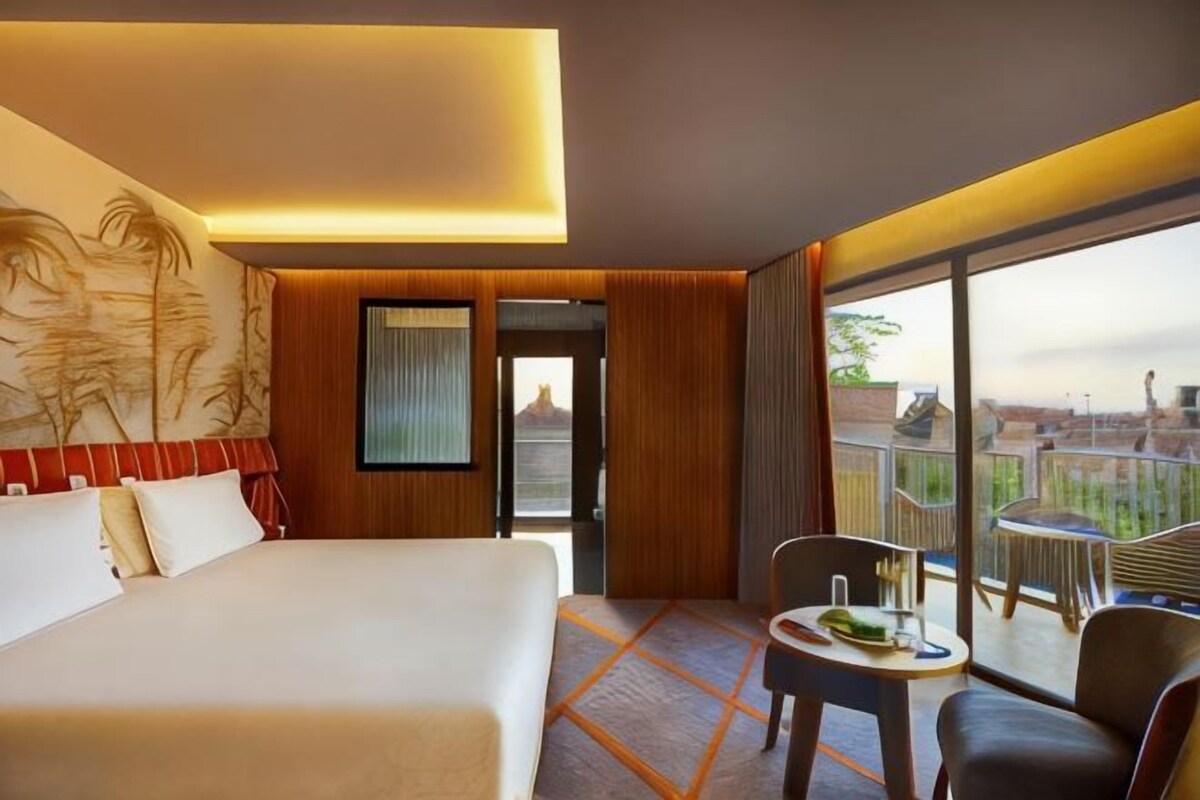 A hotel room with a large bed, a small table with a glass, a chair, and a balcony with a view.
