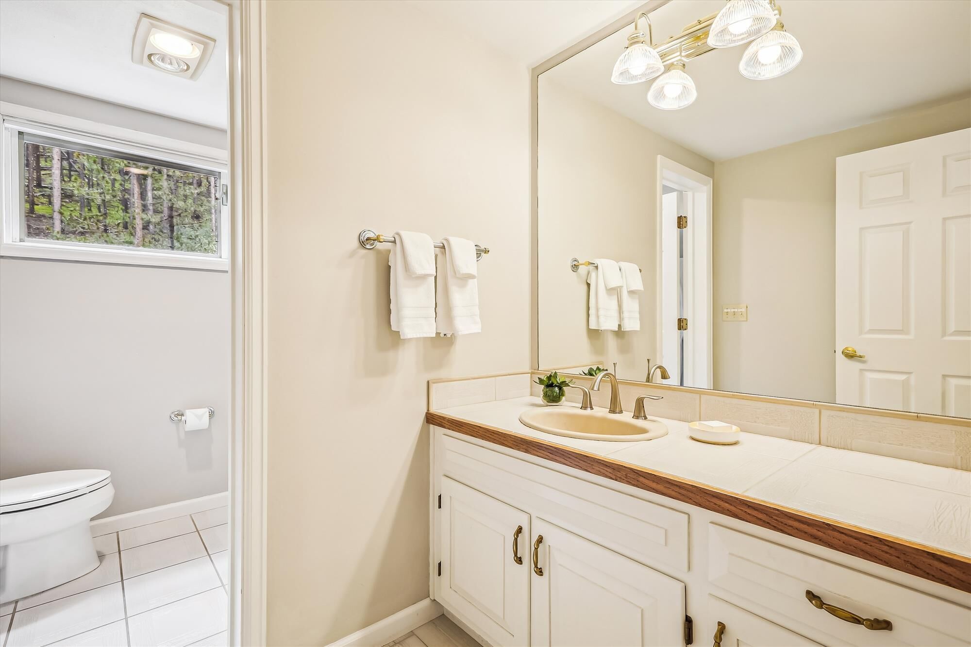 Een badkamer met een wit toilet, een wastafel met spiegel erboven en een raam met uitzicht op bomen.