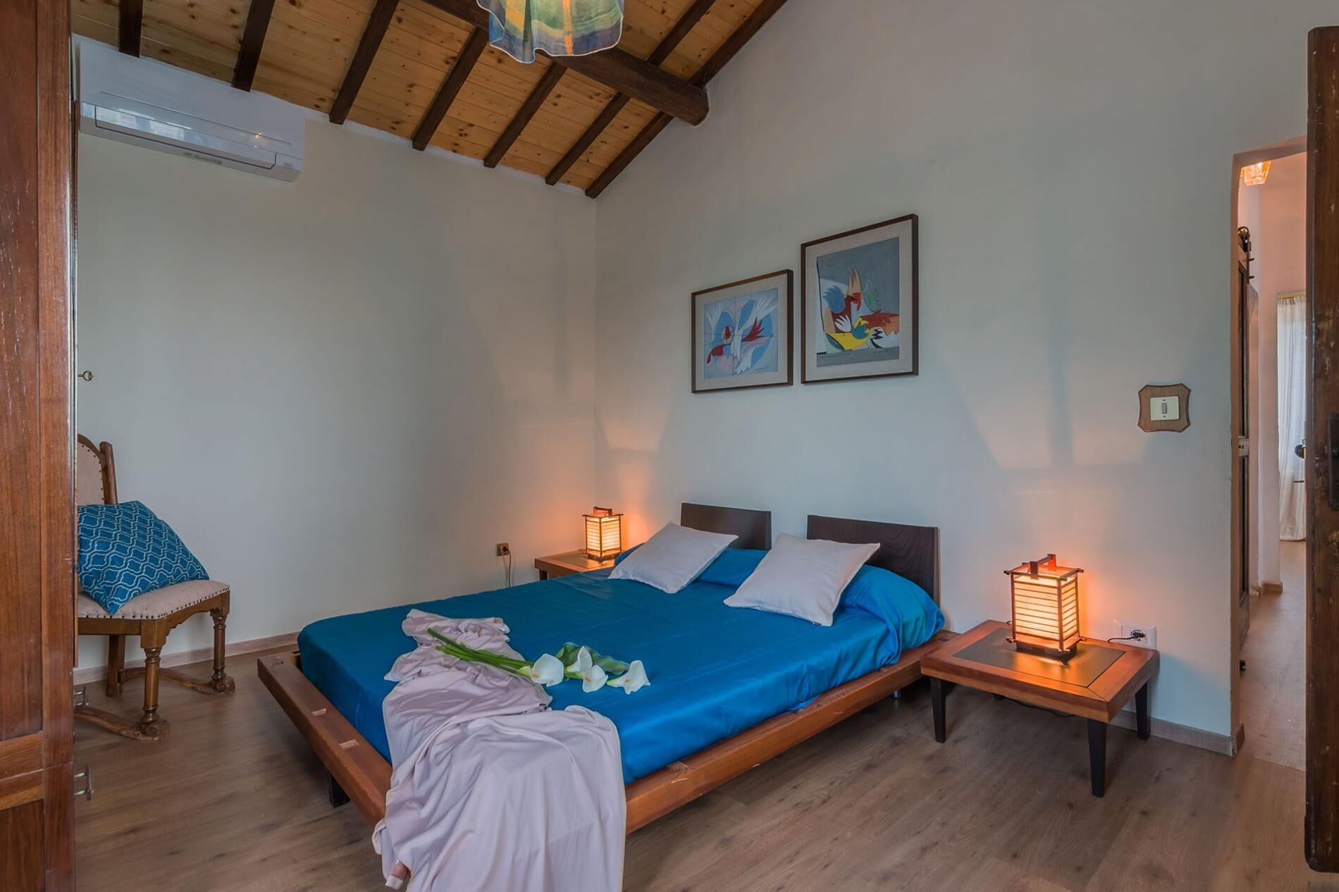 A bedroom with a wooden ceiling, a bed with blue bedding, two bedside tables with lamps, and two framed artworks on the wall.