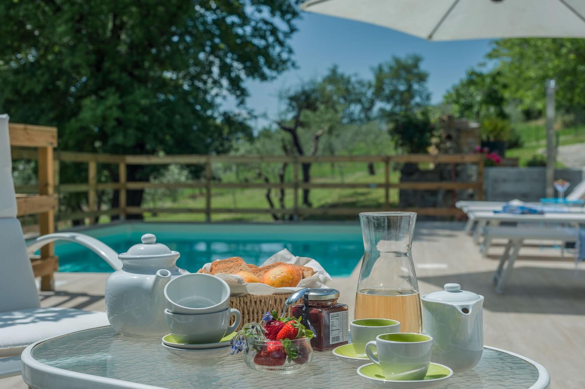 A table set with tea, a teapot, cups, a jug, and a basket of bread and strawberries by a poolside.