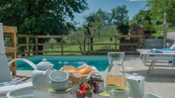 A table set with tea, a teapot, cups, a jug, and a basket of bread and strawberries by a poolside.