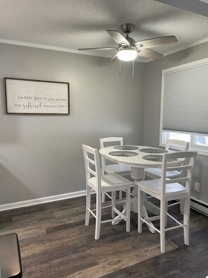 A dining area with a round table, white chairs, a ceiling fan, and a framed quote on the wall.