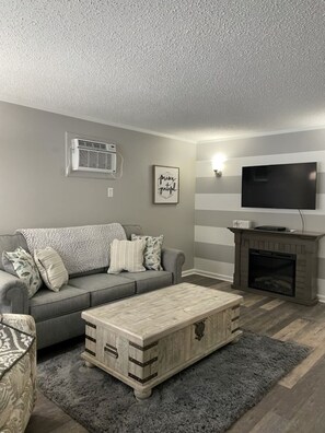 Ein Wohnzimmer mit grauem Sofa, einem Holztisch, einem Kamin, einem Wand-Fernseher und einer Klimaanlage.