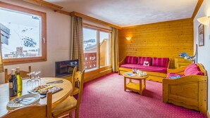 A cozy room with a wooden floor, a sofa, a dining table with plates and glasses, and a window with curtains. - travelski home select - Résidence Aspen 4* - 3 pièces 6/7 personnes (La Plagne)