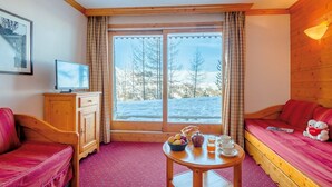 A cozy room with a wooden interior, a large window with a view of a snowy landscape, a small table with breakfast items, and a red sofa. - travelski home select - Résidence Aspen 4* - 3 pièces 6/7 personnes (La Plagne)