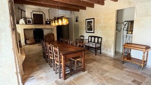 Een ruime eethoek met een grote houten tafel en stoelen, een hanglamp en een vloer van stenen tegels.