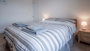 Een net opgemaakt bed met gestreept beddengoed, een bedlampje en opgevouwen handdoeken op het nachtkastje.