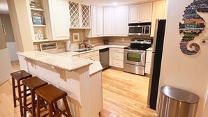 A kitchen with white cabinets, a stainless steel refrigerator, a microwave, and a sink. There are wooden stools at the island. - Tybee Retreat Unit 2 (Tybee Island)