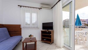 Et værelse med en sofa, et fjernsyn på et træstel, et lille bord med en plante, og en balkon med en blå parasol.