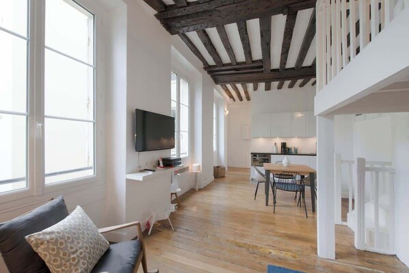 A spacious living area with a wooden floor, a sofa with patterned cushions, a television, and a kitchen in the background.