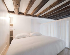 Quarto com cama, paredes brancas e vigamento de madeira no teto.