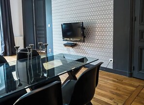 Uma área de refeições moderna com mesa de vidro e cadeiras pretas, uma TV fixada na parede e papel de parede estampado.