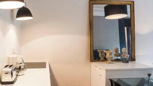 A modern kitchenette with a sink, a coffee maker, and a toaster. There is a mirror with a gold frame and a black lamp on the wall.