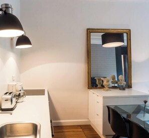 Quarto de hotel compacto com pia branca, uma pequena mesa, espelho com moldura dourada e duas luminárias fixadas na parede.