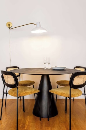 Uma mesa de jantar redonda com quatro cadeiras, uma luminária pendente branca e dois copos de vinho sobre a mesa.