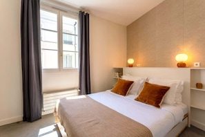 Quarto com cama, janela com cortinas, mesa de cabeceira com abajur e prateleira na parede.