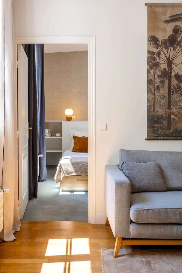 A modern living room with a grey sofa, a wooden floor, and a doorway leading to a bedroom with a bed and a nightstand.