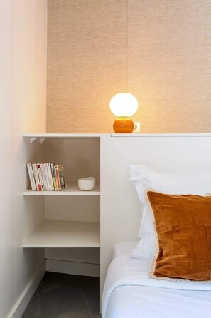 A neatly arranged bedroom with a white bed, a brown throw pillow, a shelf with books, and a wall-mounted lamp.