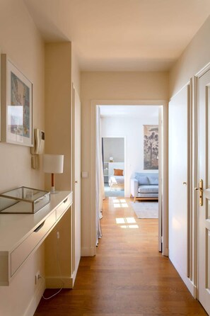 A hallway leading to a bedroom with a bed, a bedside table, and a lamp. There is a door on the right and a wall with a framed picture.