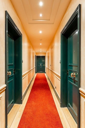 Corredor de hotel com carpete vermelho, paredes em tom de bege e portas verde-escuras.
