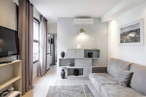 Uma sala de estar moderna com um sofá cinzento, uma televisão de ecrã plano, um console de mesa branco e uma janela grande com cortinas.