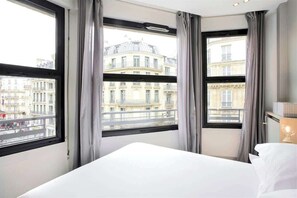 A hotel room with a large bed, two windows with curtains, and a view of Parisian buildings.