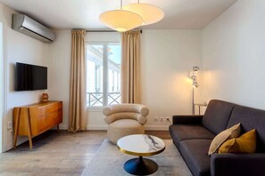 Uma sala de estar moderna com sofá, mesa de centro redonda, poltrona estofada, aparador de madeira, TV de tela plana e uma janela grande com cortinas.