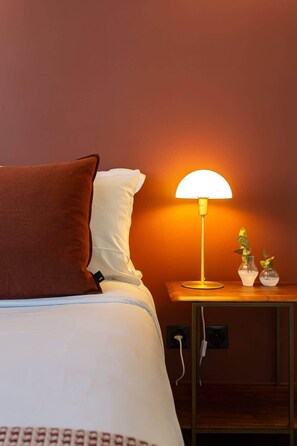 Uma cama bem arrumada com travesseiro marrom e lençóis brancos, uma luminária de cabeceira e dois pequenos vasos com plantas sobre uma mesinha de cabeceira de madeira.