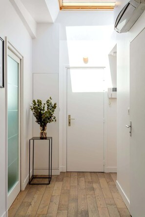 Un couloir avec une porte blanche, une petite table sur laquelle est posée une plante, et une lucarne au plafond.