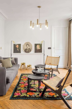 Een moderne woonkamer met een grijze bank, een ronde zwarte salontafel en een rieten fauteuil. Drie ingelijste portretten hangen aan de muur en erboven een kroonluchter.
