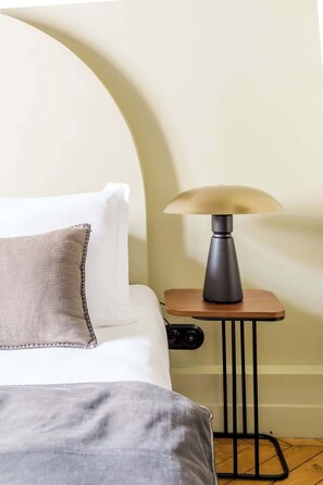 Een net opgemaakt bed met een grijze deken en een beige kussen, een klein nachtkastje met een lamp, en een houten vloer.
