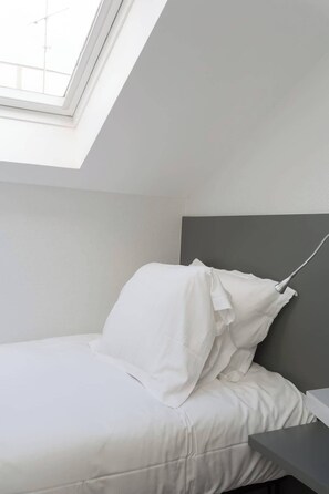 Quarto com claraboia, cama com roupa de cama branca e cabeceira cinza.