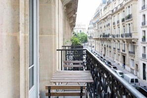 A narrow balcony with a metal railing, a wooden bench, and a potted plant.