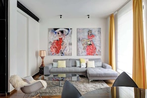 Uma sala de estar moderna com um sofá cinzento, uma mesa de centro de vidro e uma poltrona aconchegante. Há duas pinturas abstratas na parede e um abajur sobre uma mesinha lateral.