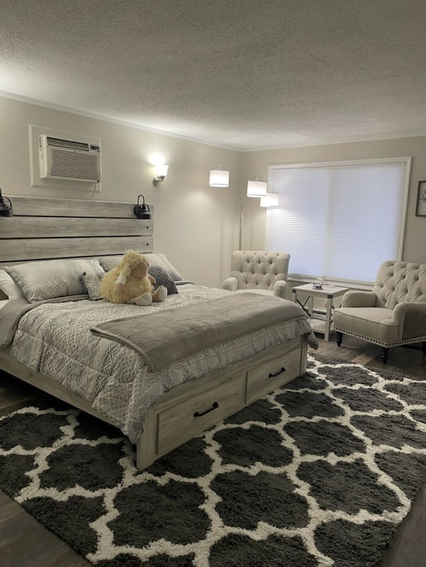 A bedroom with a bed, two chairs, a small table, a rug, and a window with blinds. - Adventure Acres- Suite C8 (Holly)
