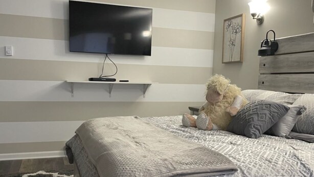 A bedroom with a bed, a TV mounted on the wall, a bedside table, and a nightstand. - Adventure Acres- Suite C8  (Holly)
