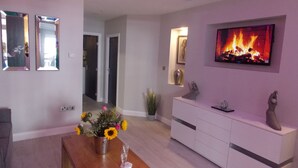 Una sala moderna con una mesa de centro de madera, un jarrón de girasoles, un televisor que muestra una chimenea y un armario blanco.