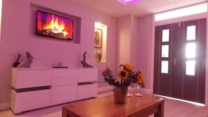 Una sala moderna con una mesa de centro de madera, mueble blanco para entretenimiento, televisor de pantalla plana mostrando una chimenea y una puerta con amplias ventanas.