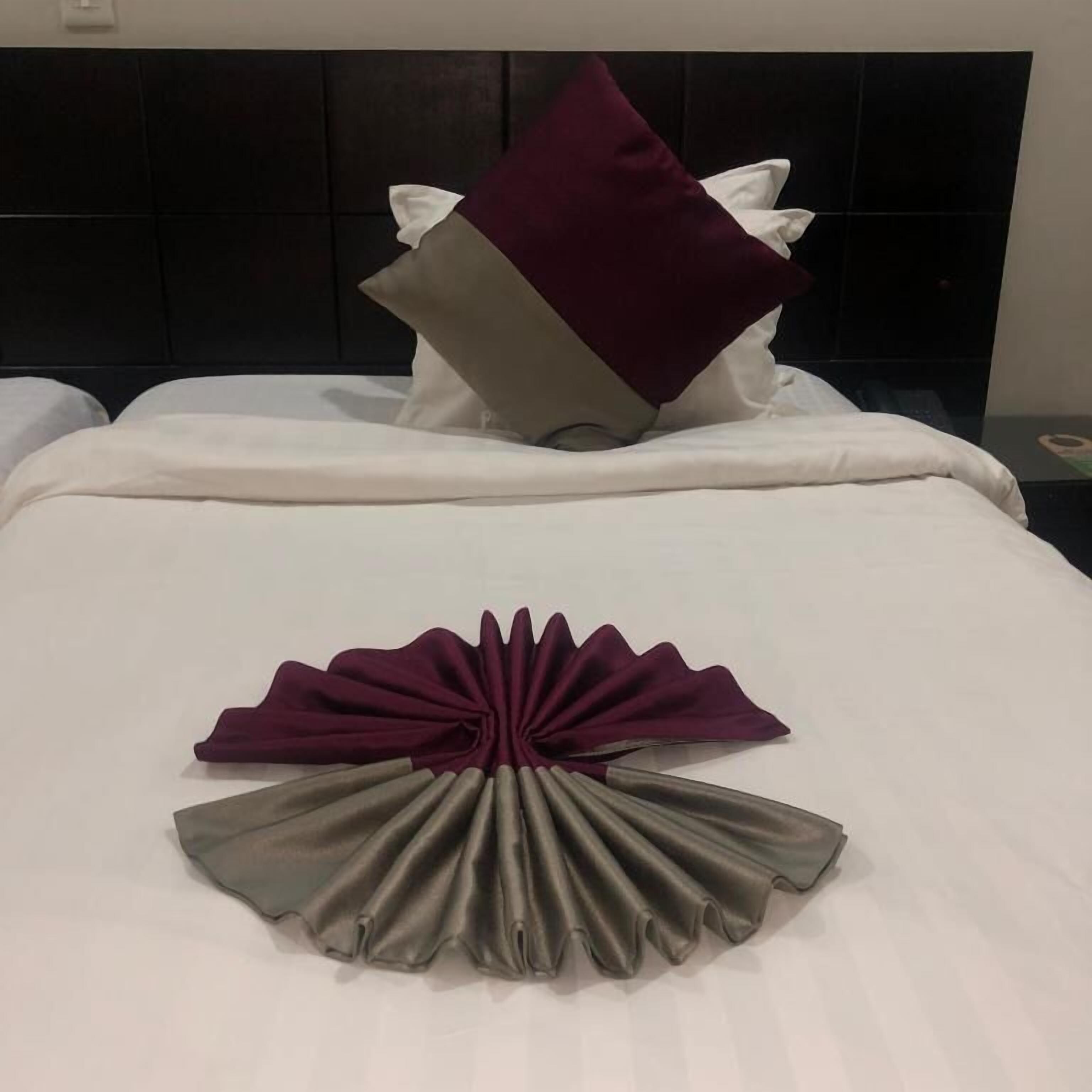 Una cama bien hecha con ropa de cama blanca, una almohada rojo oscuro y un arreglo decorativo de tela gris y morado.