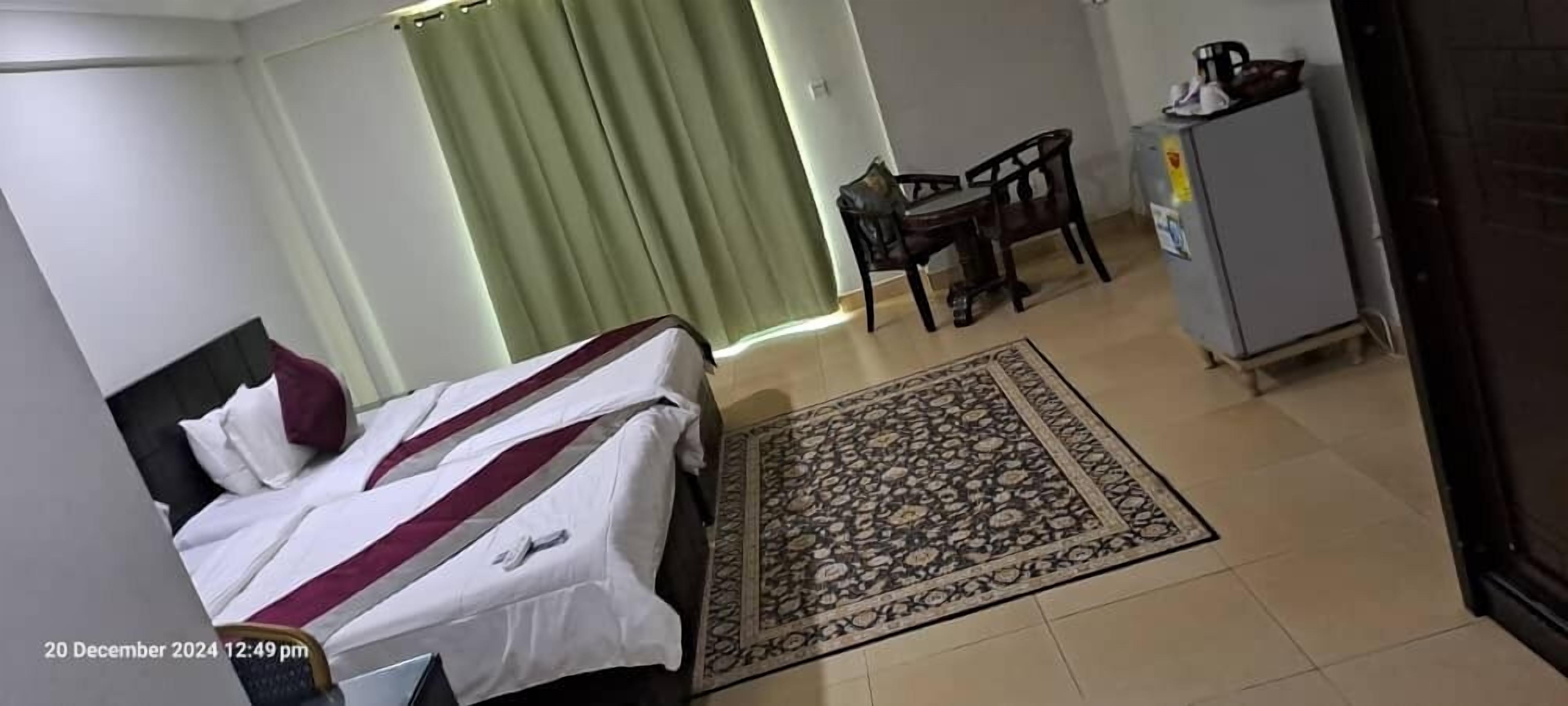 A hotel room with a bed, a desk, a chair, a refrigerator, and a patterned rug.