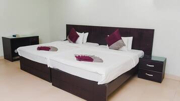 Una cama doble con cabecera oscura, ropa de cama blanca y dos almohadas decorativas. Hay una mesita de noche con una lámpara y un cajón pequeño.