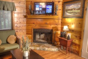A cozy living room with a fireplace, a television mounted above it, and a framed landscape painting on the wall.