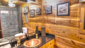 Une salle de bain rustique, située dans une cabane en bois, avec une douche en pierre, un lavabo en cuivre et des tableaux encadrés accrochés aux murs.